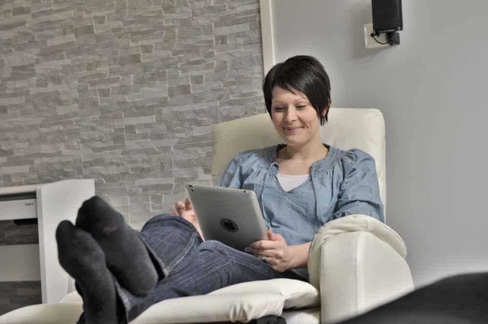Lyhythiuksinen nainen istuu nojatuolissa mukavasti jalat ylös nostettuina tablettia selailemassa. 