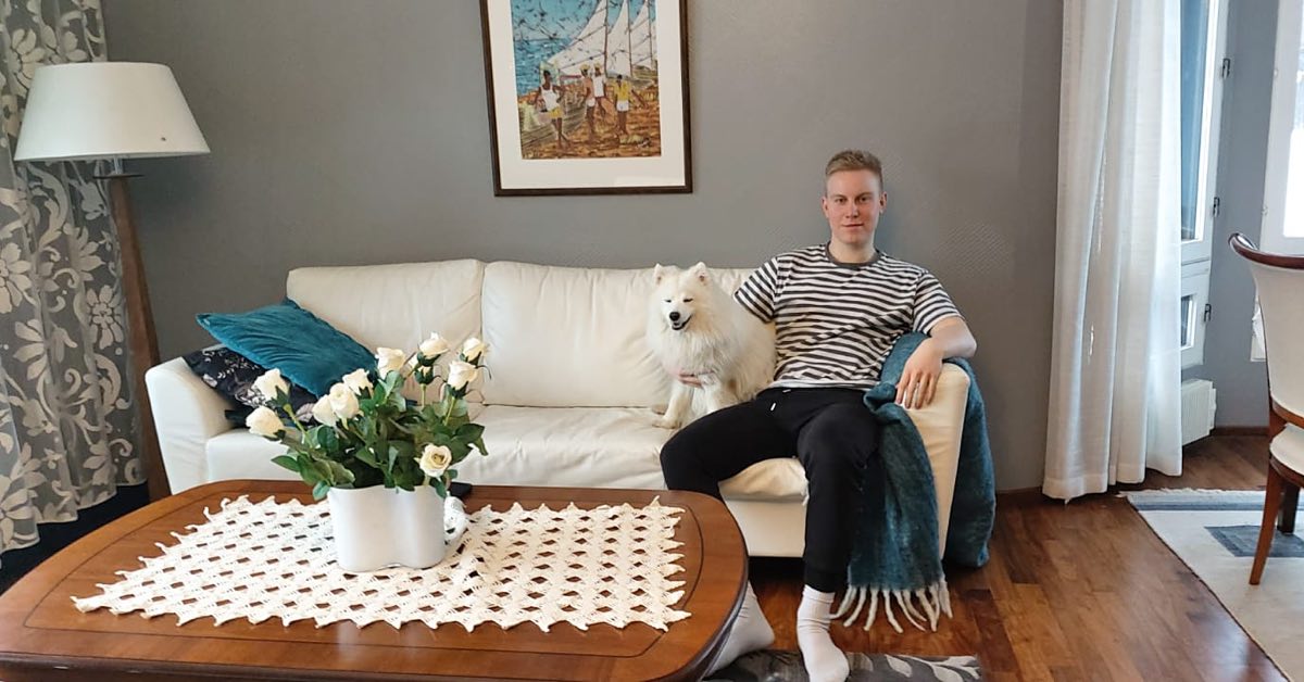 Nuori mies istuu hymyillen sohvalla vieressään pieni valkoinen koira.